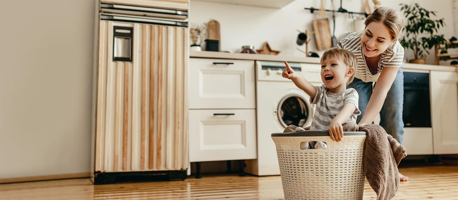 Eine Mutter schiebt spielerisch ihr Kleinkind in einem Wäschekorb über den Boden. Im Hintergrund ist eine Küchenzeile zu erkennen.